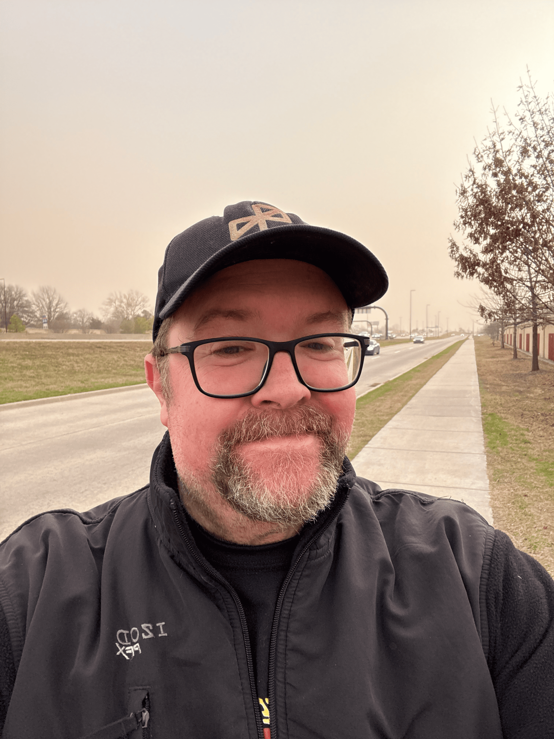 Man wearing glasses and cap taking a selfie outdoors along a roadside path under overcast sky
